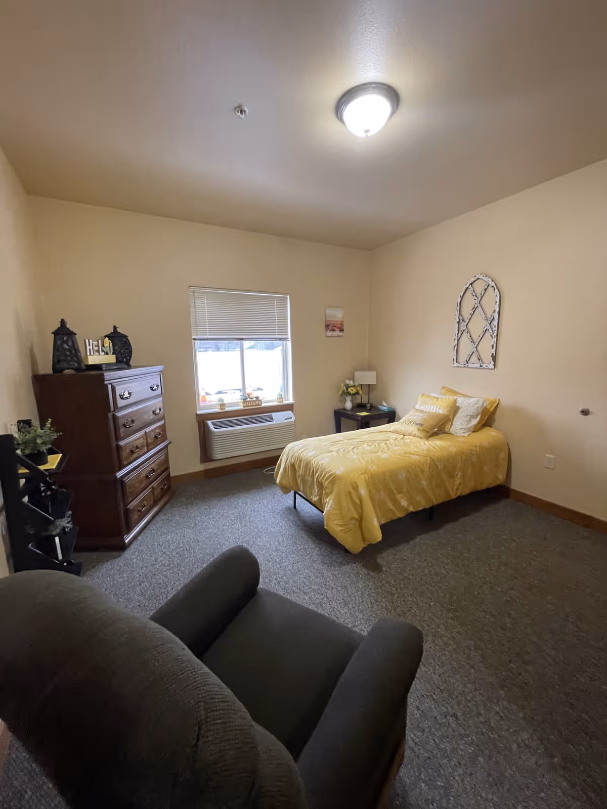 A small bedroom with a single bed covered in a yellow comforter and pillows. There is a dark armchair in the foreground, a wooden dresser with decorative items on top, a window with blinds, and a small nightstand with a lamp and flowers. The walls are beige and the floor is carpeted.