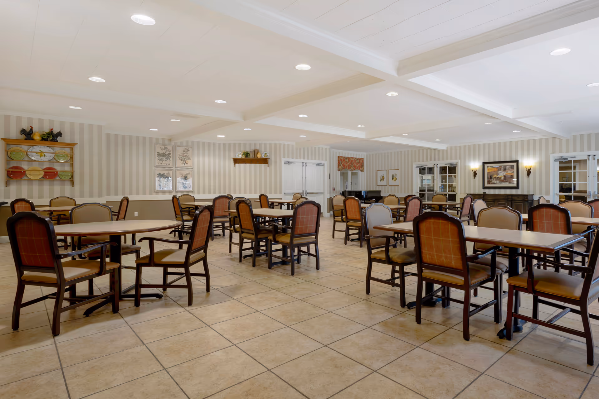 A spacious dining room with multiple tables and chairs arranged neatly. The room has tiled flooring, striped wallpaper, and recessed ceiling lights. Decorative plates and framed artwork adorn the walls, and there is a piano in the corner near double doors.