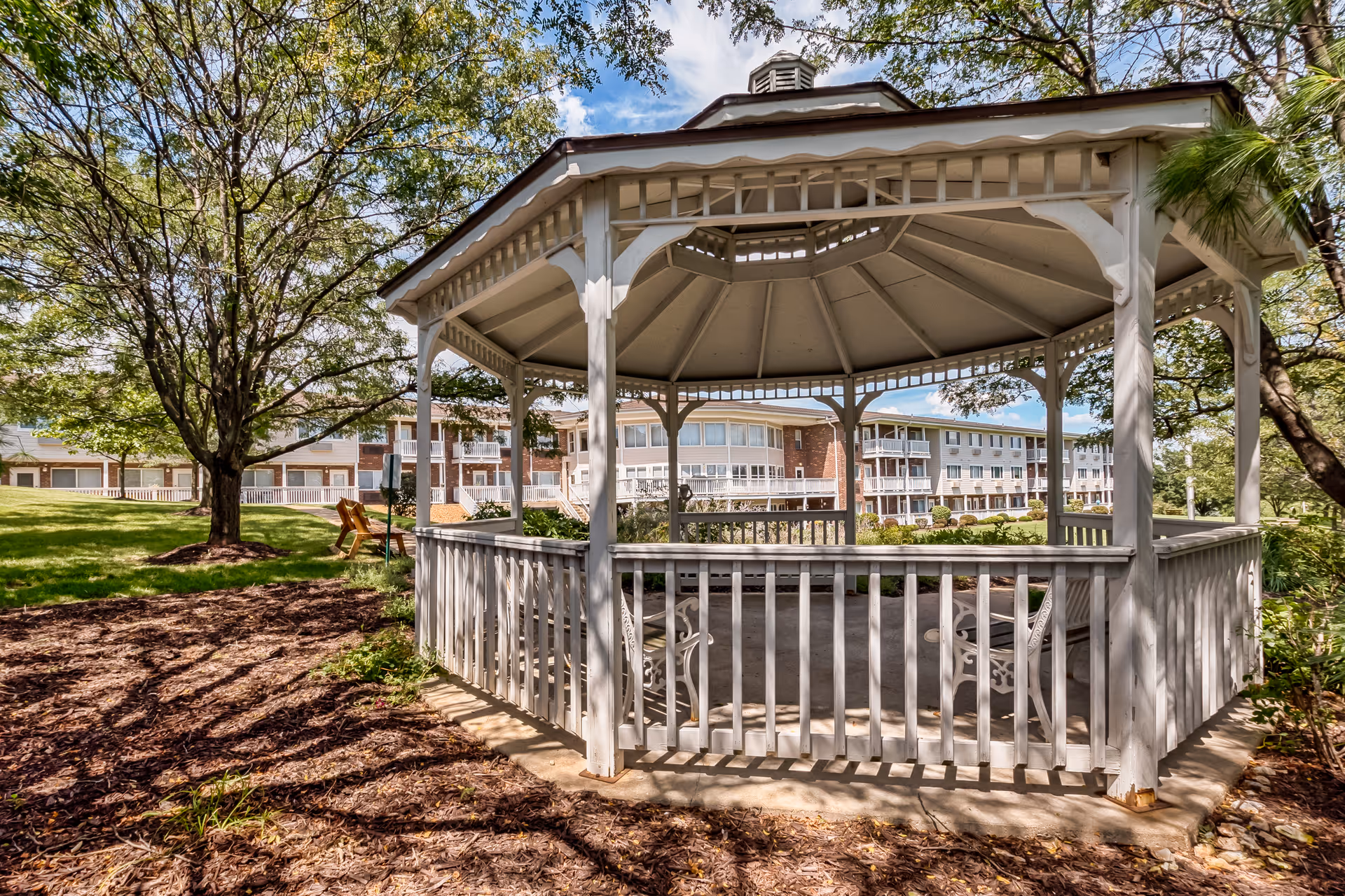 A white wooden gazebo with benches inside situated in a landscaped garden area with trees and mulch. In the background, there is a multi-story assisted living facility building under a partly cloudy sky.