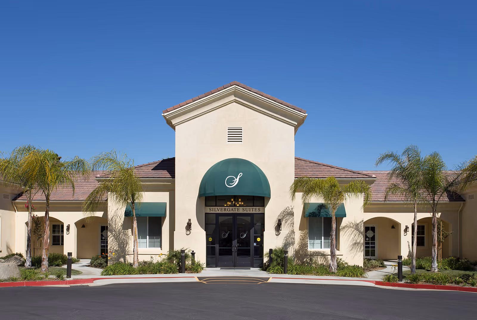 Front entrance of a beige stucco senior living building with a green awning, palm trees, and a clear blue sky.