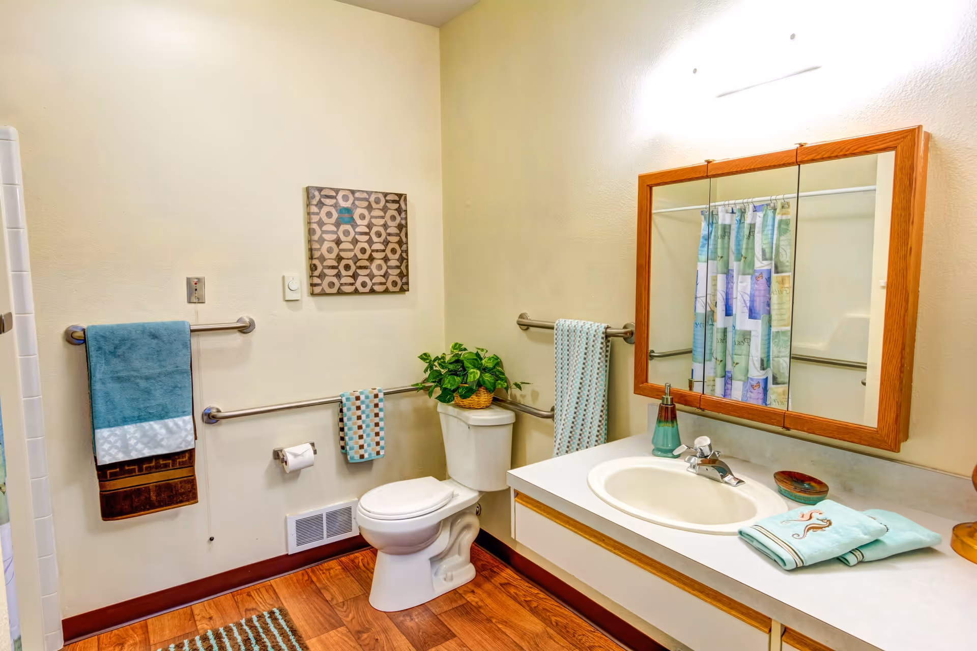 A clean and well-lit bathroom with a white toilet, a sink with a countertop, a wooden framed mirror above the sink, and a shower with a colorful curtain. There are several towels hanging on metal bars, a small plant on the toilet tank, and a decorative wall art piece. The floor has a wood-like finish and a striped rug.