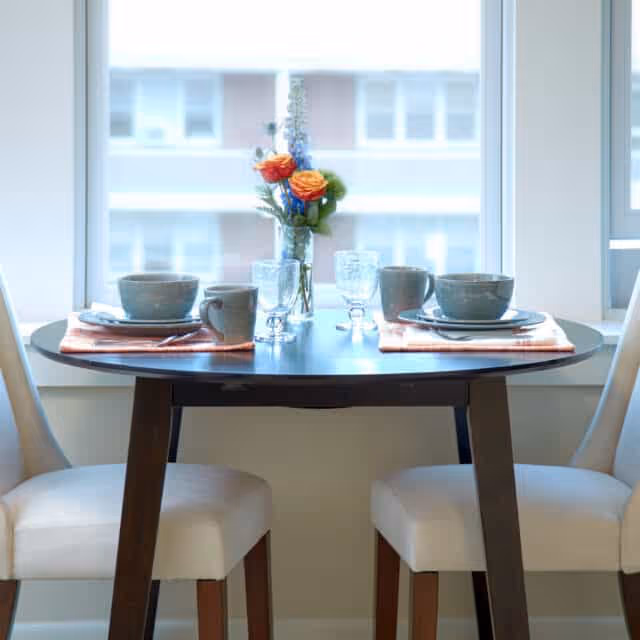 A small round dining table set for two with two white cushioned chairs. The table has two place settings with bowls, mugs, plates, and glasses on orange napkins. A small vase with orange and blue flowers is placed in the center. The background shows a large window with a blurred view of a building outside.