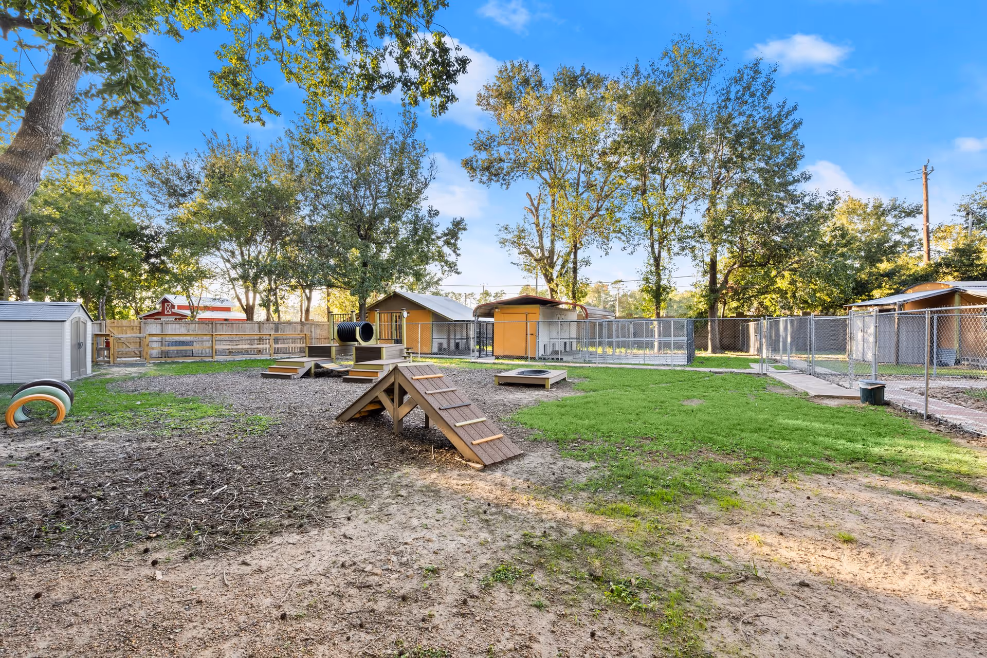 Outdoor fenced area with agility equipment including ramps, platforms, and tunnels, surrounded by trees and small sheds under a clear blue sky.