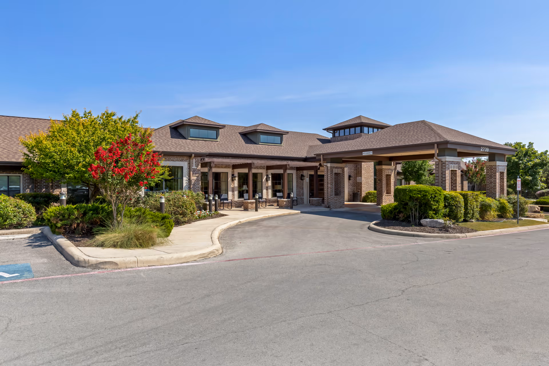 Front entrance of a single-story brick assisted living building with a covered porte-cochère, landscaped shrubs, and a clear blue sky.