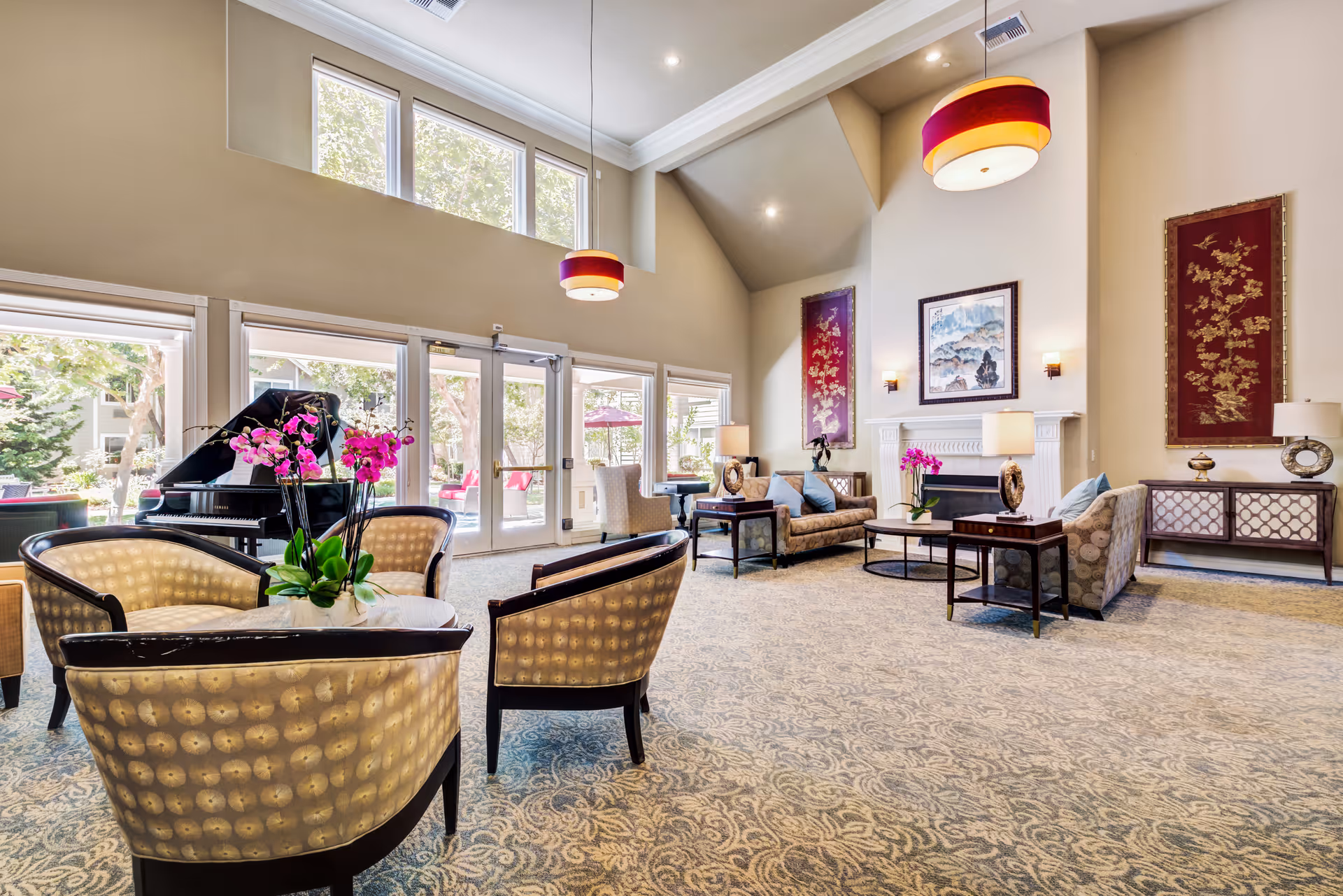 A spacious and elegant living room area with high vaulted ceilings, large windows letting in natural light, and glass doors leading outside. The room features comfortable seating including armchairs and sofas arranged around coffee tables, a grand piano, decorative wall art, and lamps. The carpet has a detailed pattern, and there are vibrant pink orchids on the tables.