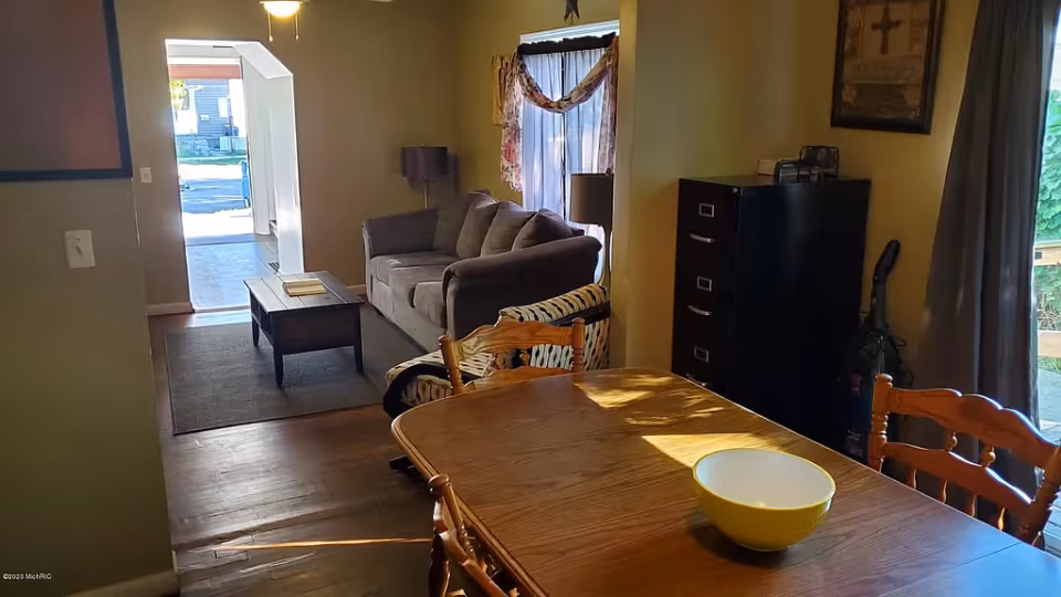 Dining table with wooden chairs and a yellow bowl in the foreground and a living room with a sofa and coffee table visible beyond.