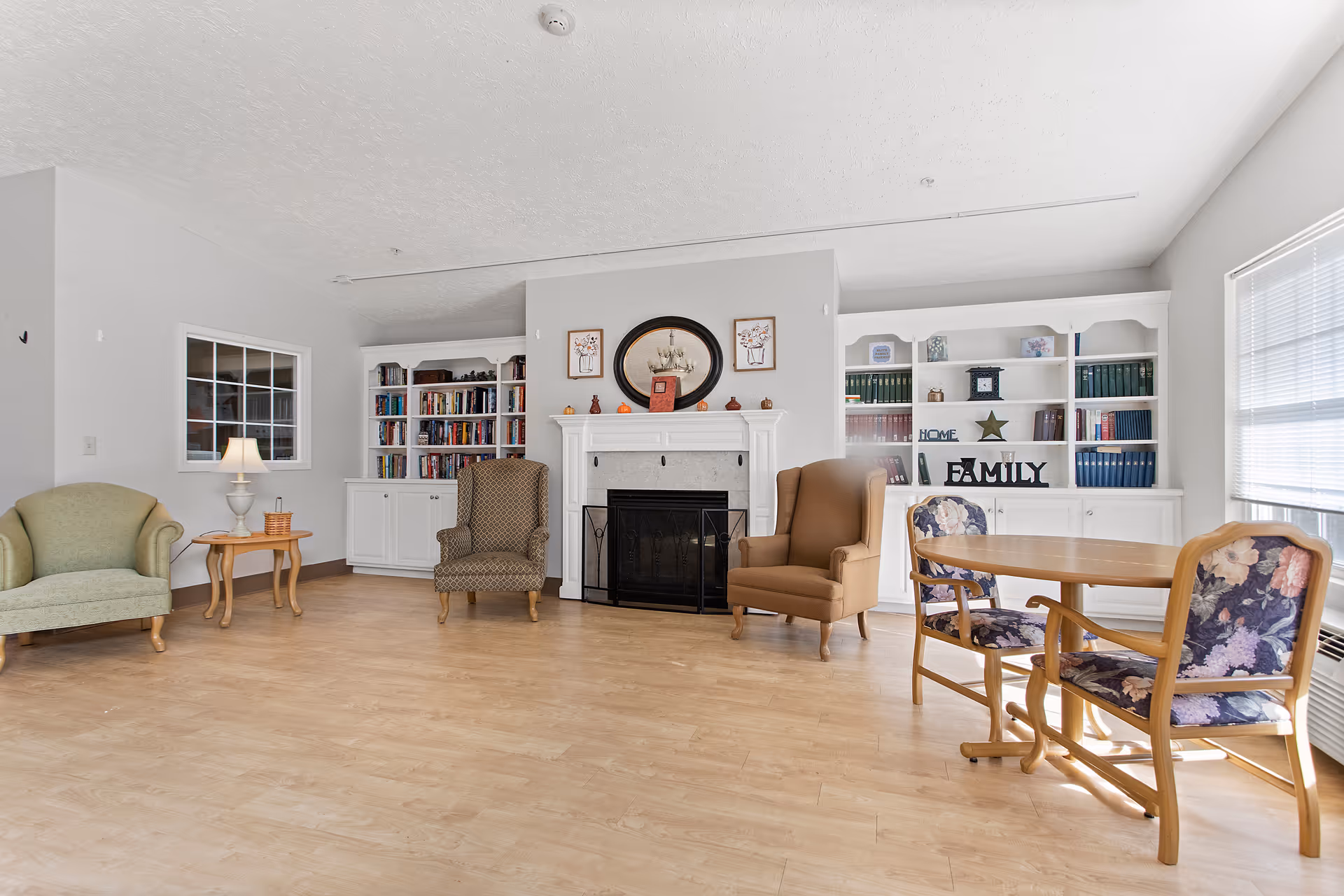 A bright and spacious living room area with light wood flooring, a white fireplace centered on the back wall flanked by built-in white bookshelves filled with books and decorative items. There are three armchairs of different styles and colors arranged around the fireplace, a small wooden side table with a lamp, and a wooden dining table with two floral upholstered chairs near a window with blinds.