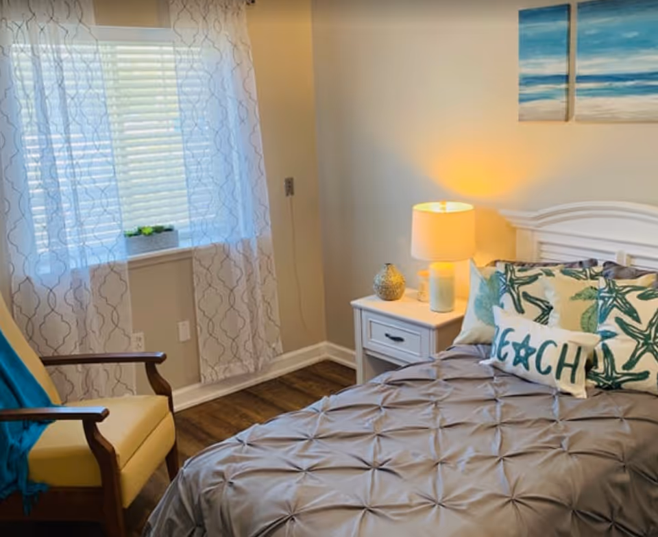 A cozy bedroom with a neatly made bed featuring a gray comforter and decorative pillows, including one with the word 'BEACH'. Next to the bed is a white nightstand with a lit lamp and decorative items. A yellow armchair with wooden arms and a blue throw blanket is positioned near a window with sheer patterned curtains. The walls are light-colored, and there are two beach-themed paintings above the bed.