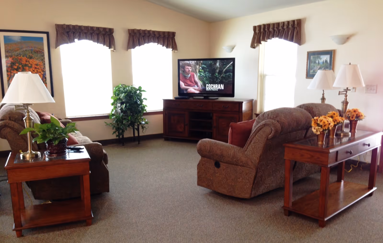 A cozy living room with two patterned sofas facing a flat-screen TV on a wooden cabinet. The room has three windows with brown valances, two wooden side tables with lamps and plants, a wooden console table with three flower vases, and framed artwork on the walls.