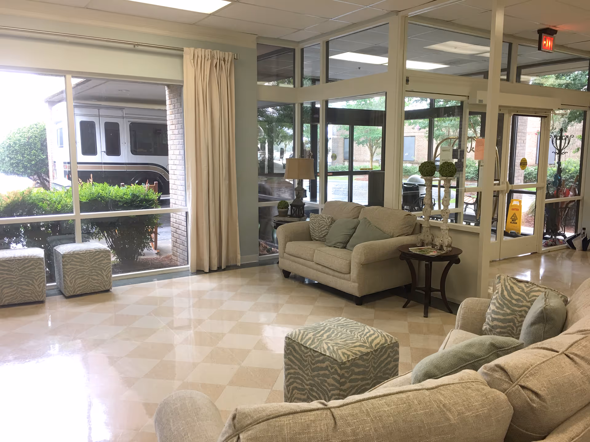 A bright and clean seating area in a healthcare facility with beige sofas, patterned ottomans, and a small wooden side table with decorative items. Large windows and glass doors provide a view of greenery and a parked vehicle outside. The floor has a checkered tile pattern, and there is an exit sign above the door.