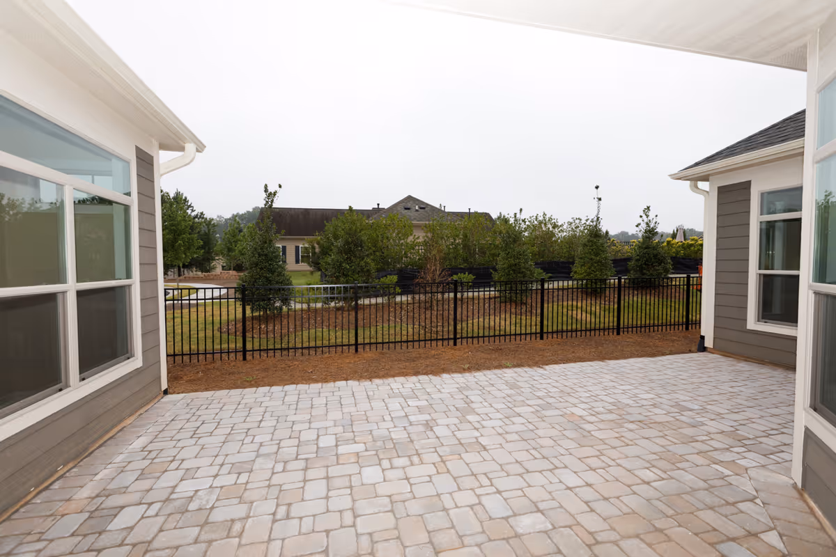 A paved outdoor patio area between two buildings with large windows, overlooking a fenced garden with small trees and shrubs under an overcast sky.
