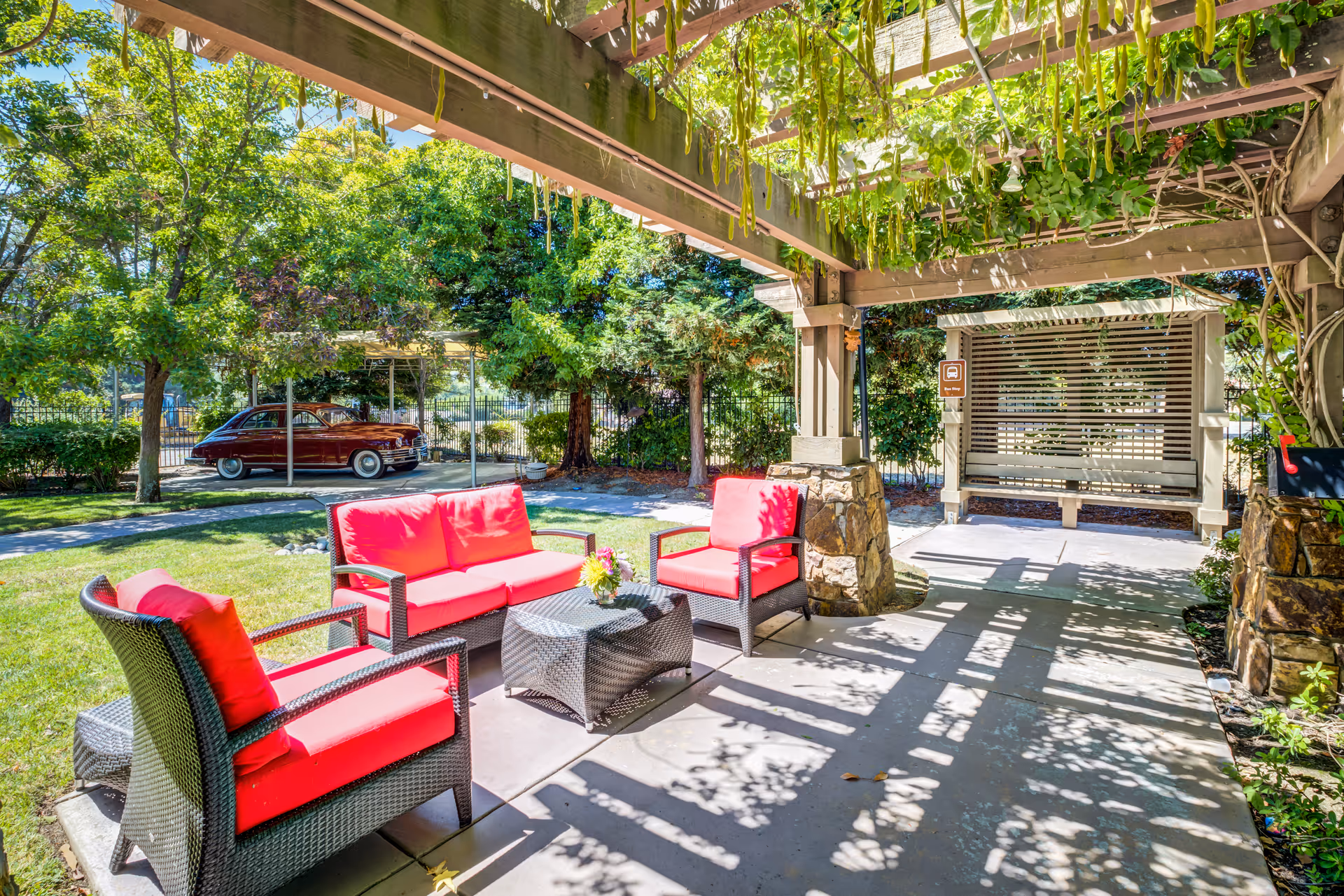 Outdoor seating area with red cushioned wicker chairs and a loveseat arranged around a wicker coffee table under a wooden pergola with hanging greenery. In the background, there is a covered bench swing and a vintage red car parked near trees and bushes.