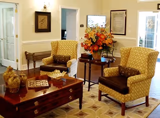A cozy living room area with two patterned yellow armchairs, a wooden coffee table with decorative items, a side table with a large floral arrangement, and framed artwork on the walls. The room has light-colored walls and a patterned rug on the floor.