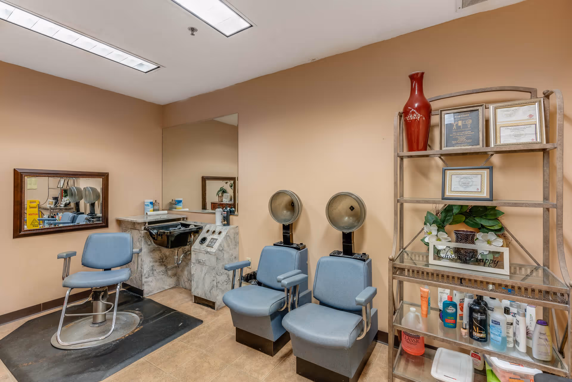 Small salon room with styling chairs and hooded hairdryers, a sink and mirror, and shelving stocked with hair products and decor.