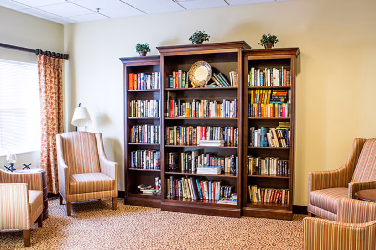 Cozy communal sitting area featuring a large wooden bookshelf filled with books flanked by striped armchairs, a floor lamp, and a window with patterned curtains.