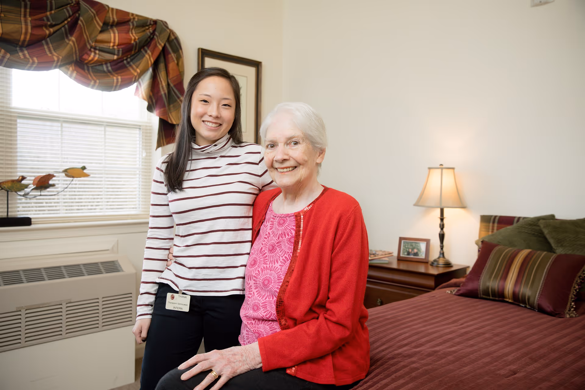 An elderly woman wearing a red cardigan and pink top sits on a bed in a cozy bedroom. A younger woman wearing a striped shirt and black pants stands beside her, smiling. The room has a window with plaid curtains, a bedside table with a lamp and framed photo, and decorative pillows on the bed.