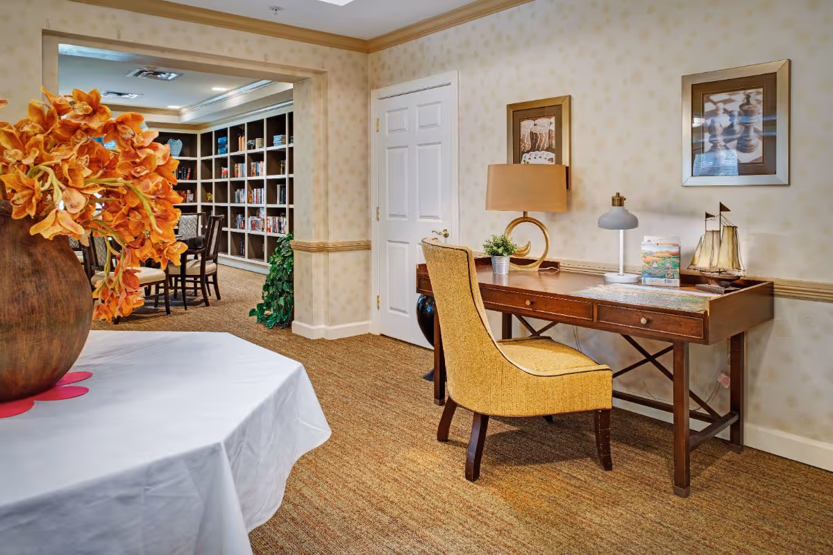Cozy interior common room with a wooden desk and yellow chair in the foreground and book-lined shelves with a table visible through an open doorway.