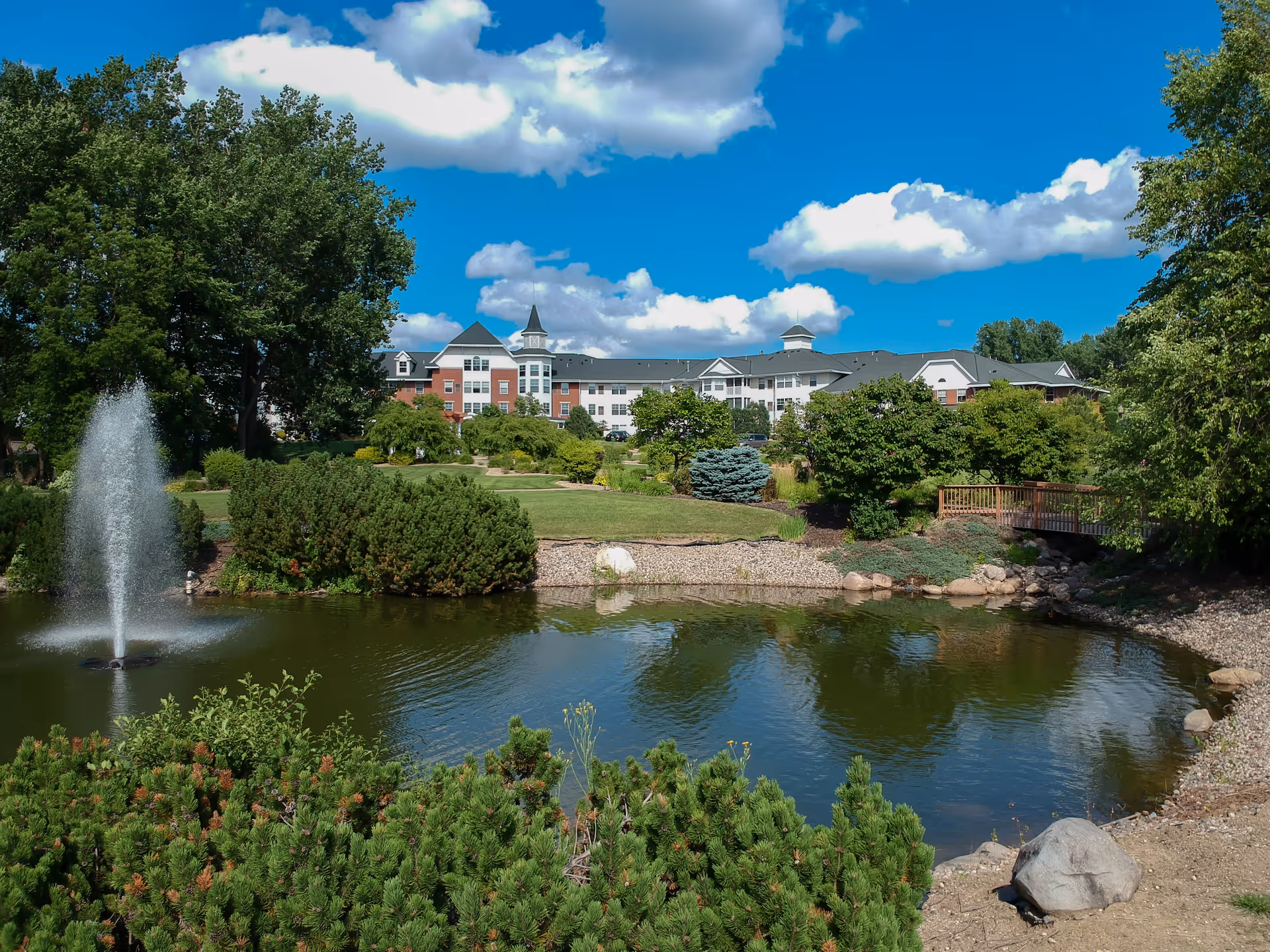 A scenic outdoor view of The Rivers Retirement Community featuring a large pond with a water fountain, surrounded by lush greenery, trees, and shrubs. In the background, there is a multi-story building with a distinctive tower and a clear blue sky with scattered white clouds.