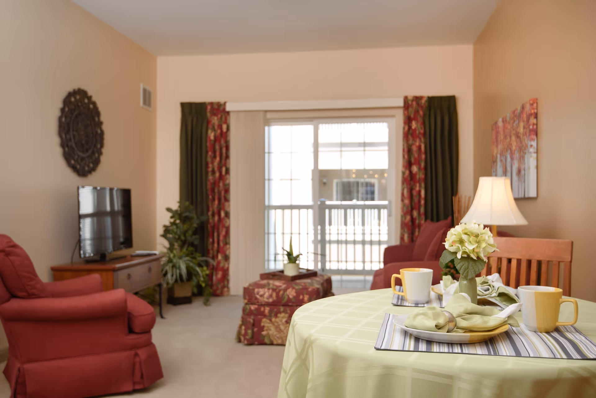 A cozy living room with a round table covered with a light green tablecloth, set with two yellow mugs, plates, and napkins. There is a small vase with flowers on the table. In the background, there are two red armchairs, a wooden TV stand with a flat-screen TV, a potted plant, and a sliding glass door with floral and green curtains.