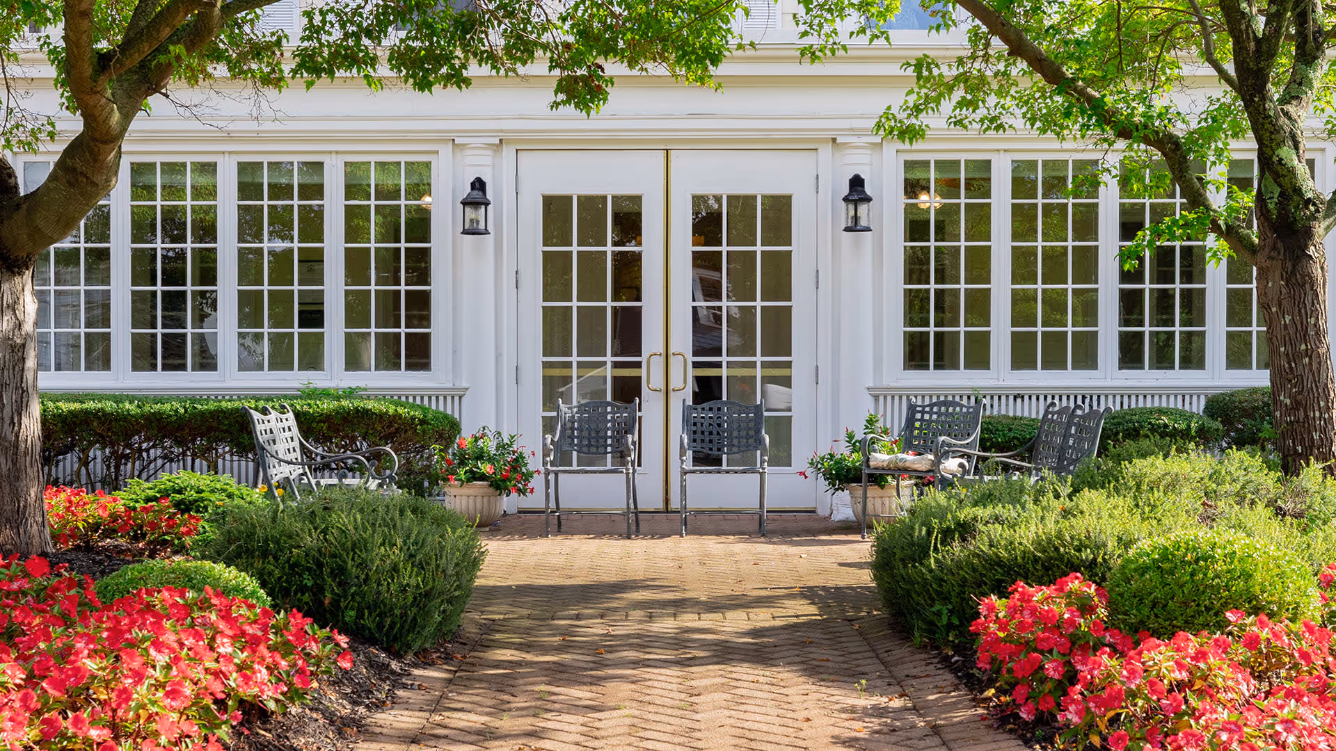 A well-maintained outdoor garden area with vibrant red flowers, green shrubs, and trees leading to a white building with large windows and double glass doors. Several metal chairs are arranged near the entrance, creating a welcoming seating area.