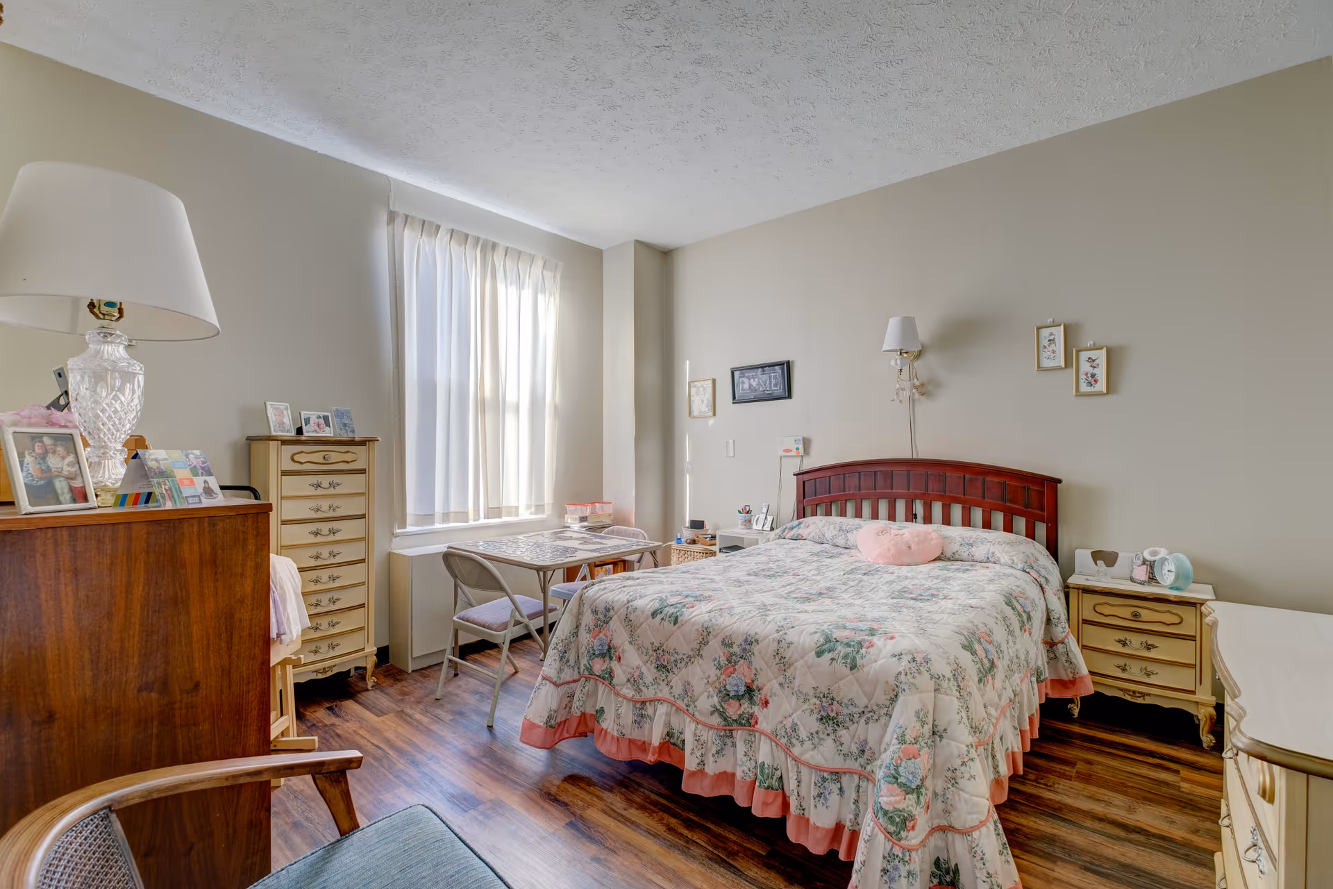 A cozy bedroom with a floral bedspread on a wooden bed frame, a small table with two chairs near a window with sheer curtains, a wooden dresser with a lamp and framed photos, and two nightstands with various items. The room has wooden flooring and light-colored walls with a few framed pictures hanging.