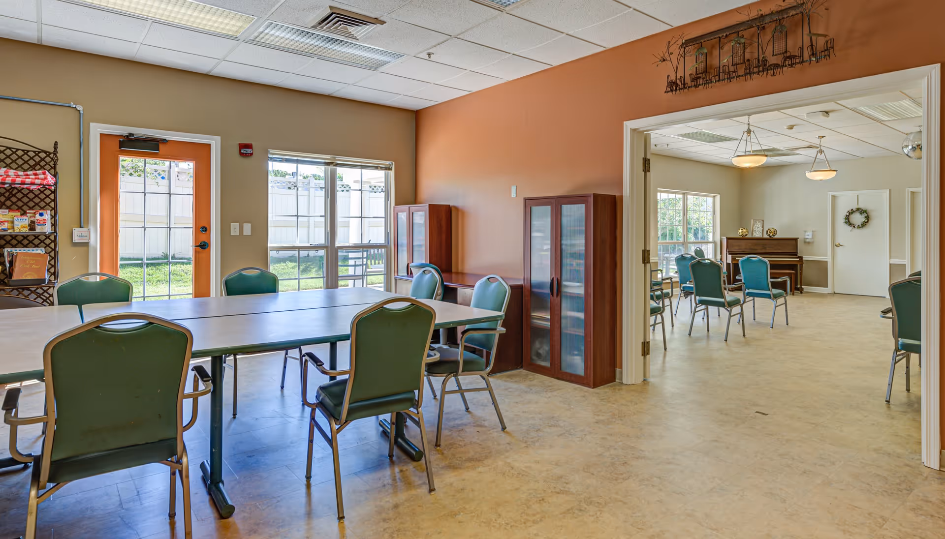 Interior view of a senior living facility showing two connected rooms with tables and green chairs. The room in the foreground has a long table with several chairs around it, a door with a window leading outside, and a shelving unit with snacks. The adjacent room has more chairs arranged around tables, a piano against the far wall, and large windows letting in natural light.