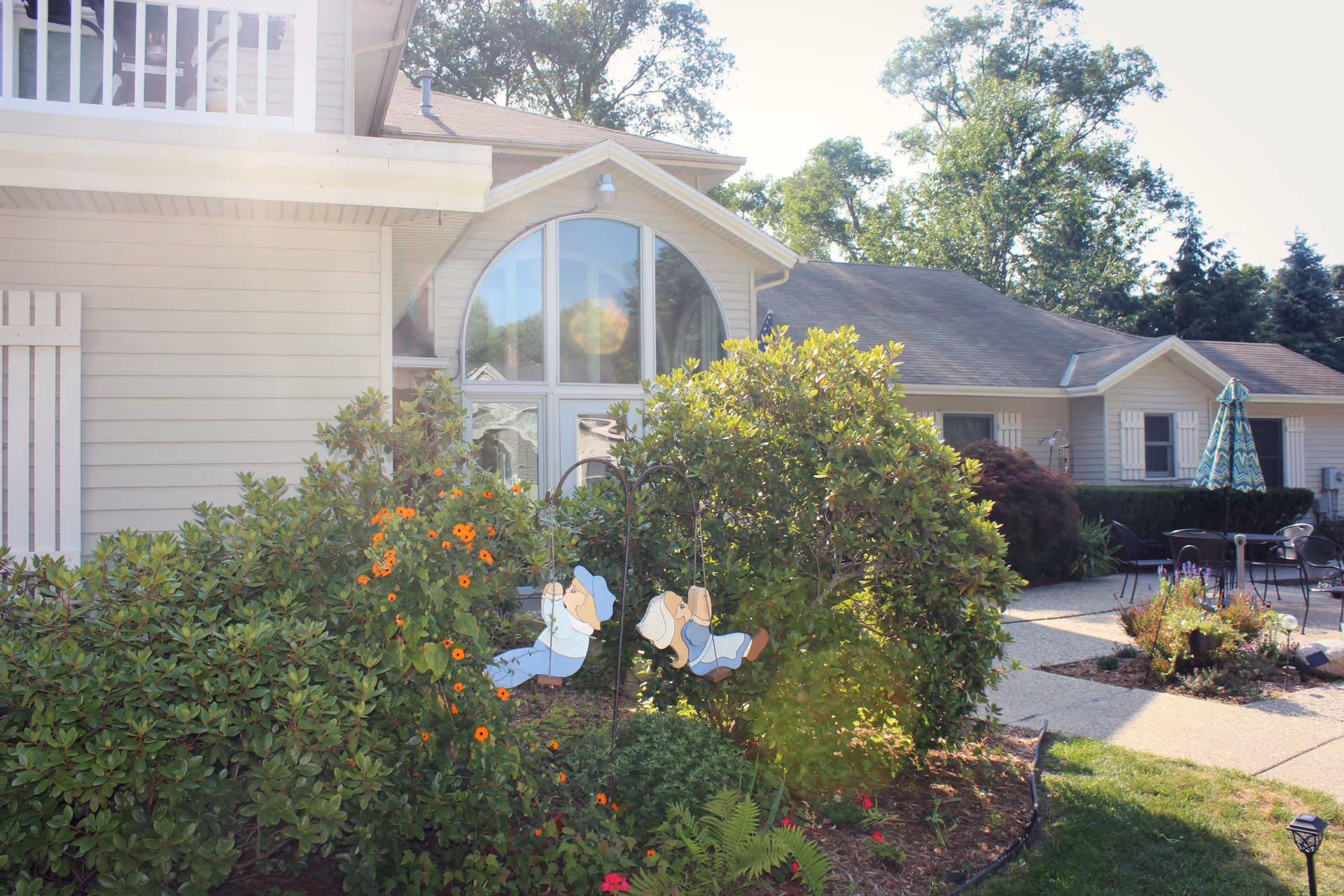 Outdoor garden area with green bushes, orange flowers, and decorative figures of children on swings. In the background, there is a light-colored building with large arched windows and a patio area with tables, chairs, and an umbrella.