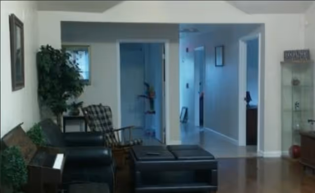 Interior view of a living room area with a black leather sofa, a wooden rocking chair with a plaid cushion, a piano on the left, a small table with a plant, and a glass display cabinet on the right. The room has white walls and a hallway leading to other rooms in the background.