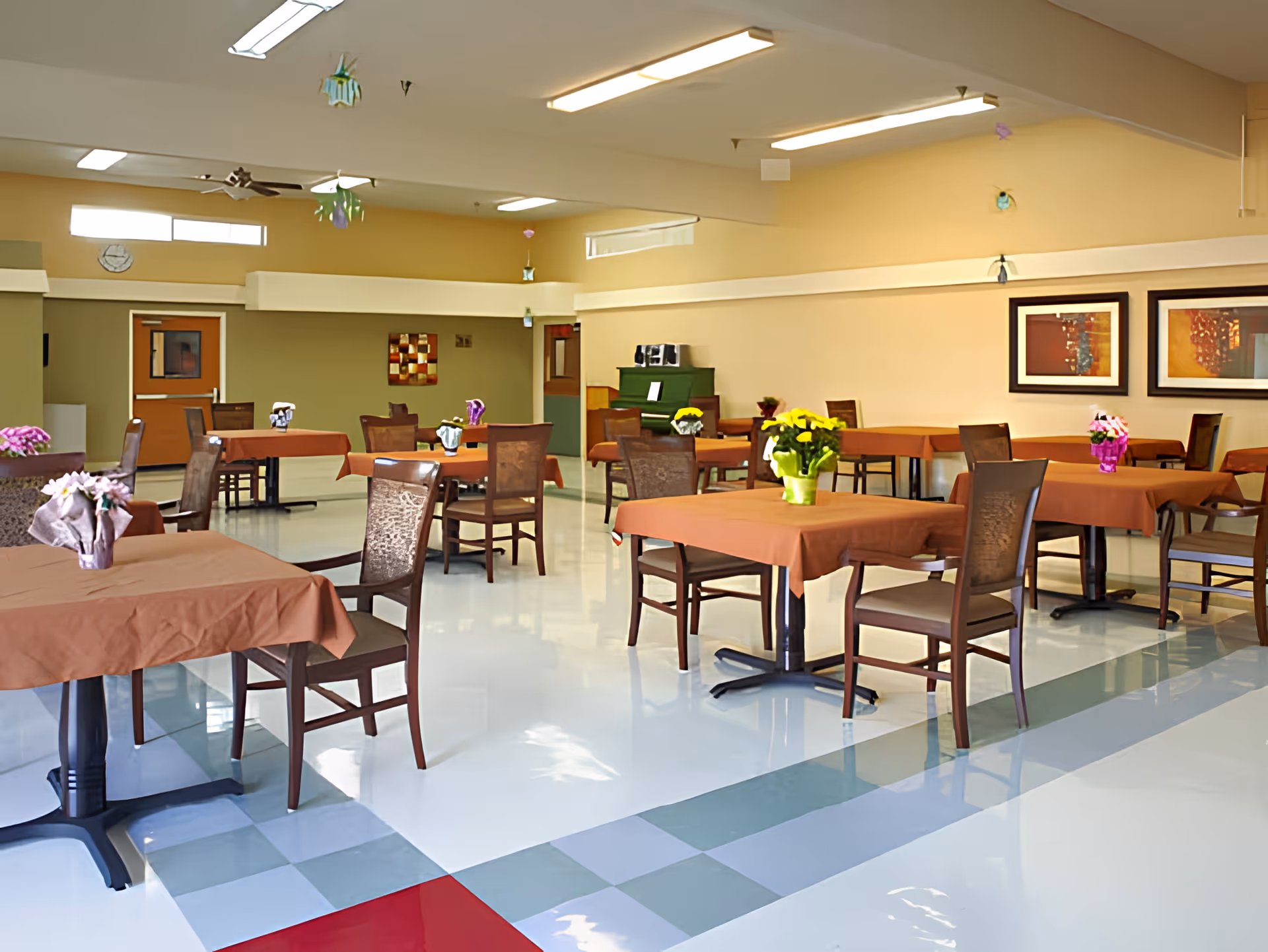 A spacious dining room with multiple tables covered in brown tablecloths, each adorned with colorful flower arrangements. Wooden chairs are placed around the tables. The room has a polished floor with a checkered pattern near the entrance, beige walls, ceiling lights, and framed artwork on the walls. A green piano and a clock are visible in the background.