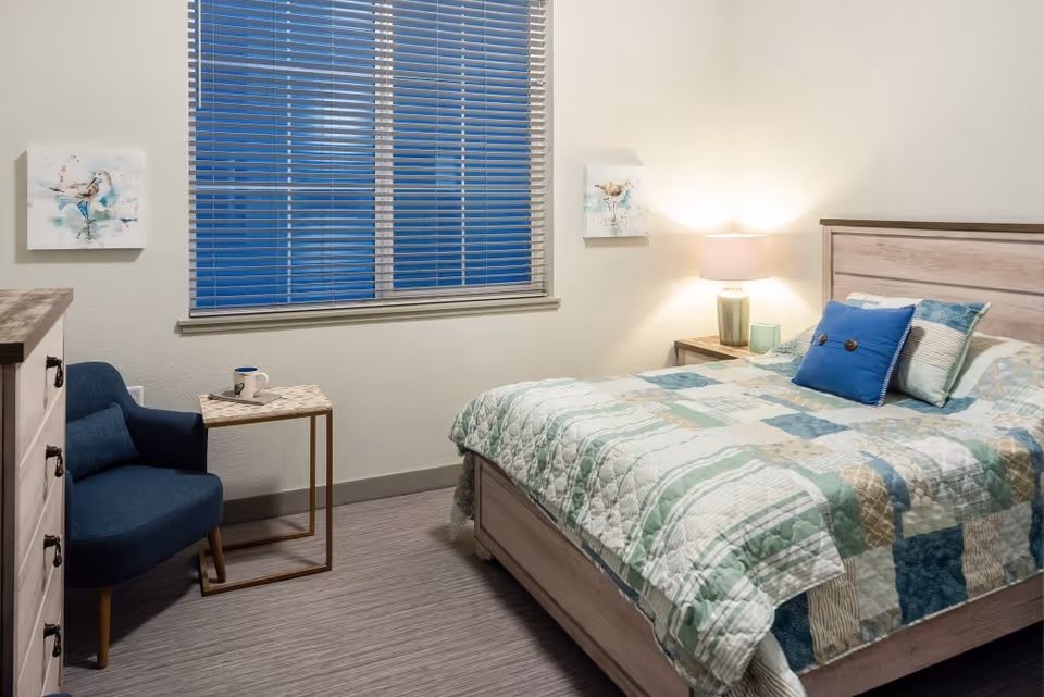 Well-lit bedroom with a bed dressed in a patchwork quilt, a bedside table and lamp, a blue armchair, and a dresser beneath a window with blinds.