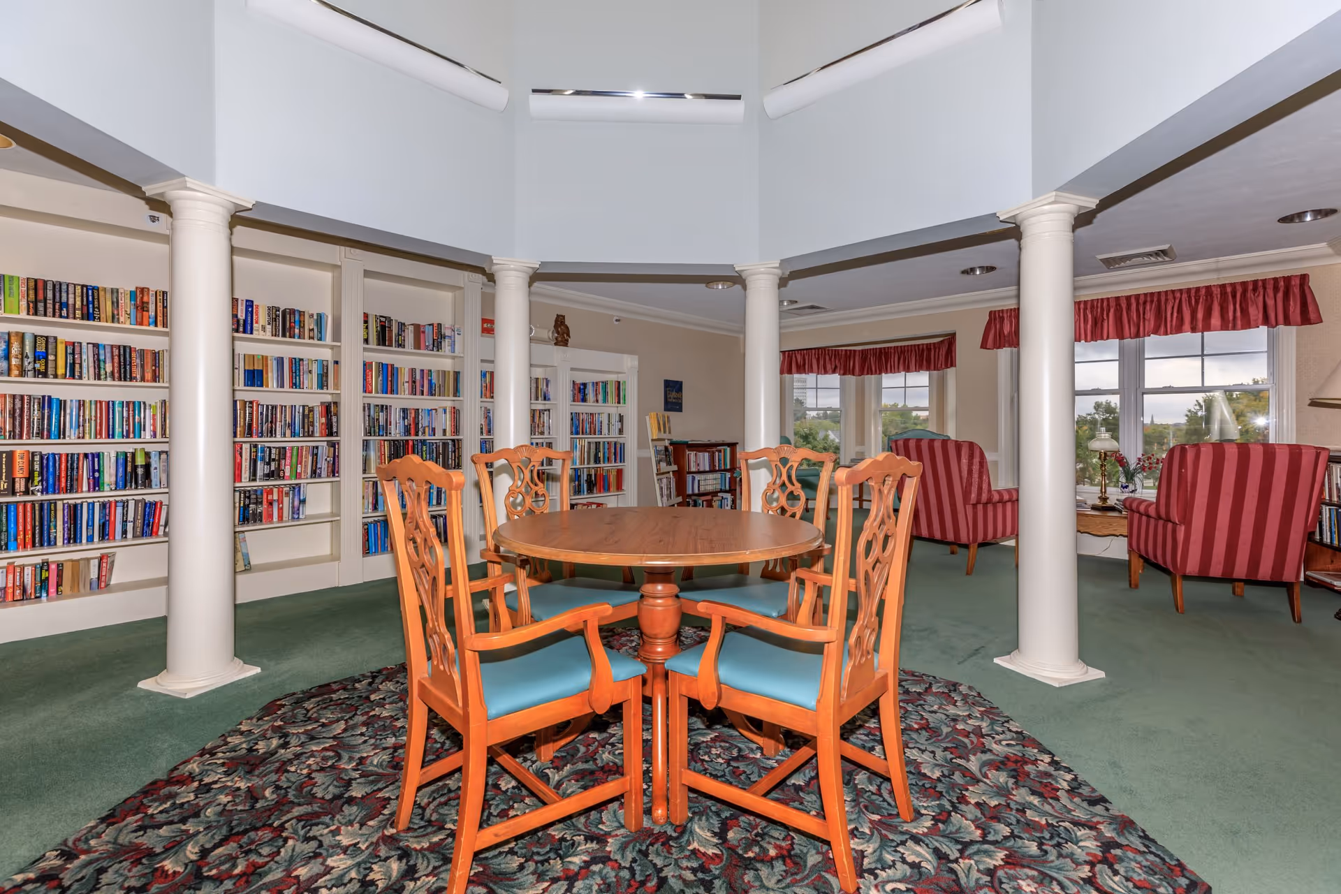A cozy library and sitting area in a senior living facility with a round wooden table surrounded by four wooden chairs with blue cushions on a patterned rug. The room features white columns, bookshelves filled with books, green carpet, and two red striped armchairs near windows with red valances.