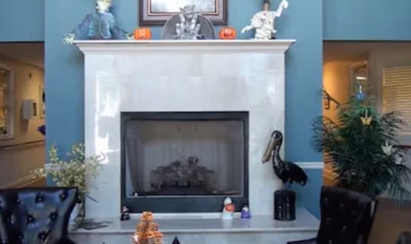 A cozy living room area featuring a white marble fireplace with Halloween decorations on the mantel, including small pumpkins and figurines of a mummy, a bat, and a Frankenstein monster. There are two black leather chairs on either side of the fireplace, a large green plant with butterfly decorations on the right, and a hallway visible in the background.
