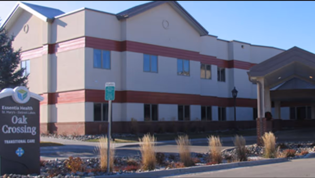 Exterior view of Essentia Health Oak Crossing building with a sign in front indicating it provides transitional care. The building is two stories with beige walls and red horizontal accents, surrounded by landscaping and a driveway.