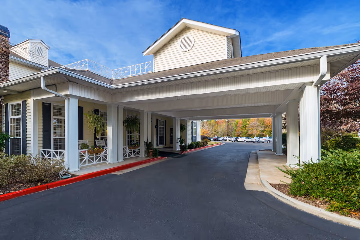 Covered driveway entrance to a senior living facility with white columns and beige siding, surrounded by greenery and a parking lot in the background under a blue sky.