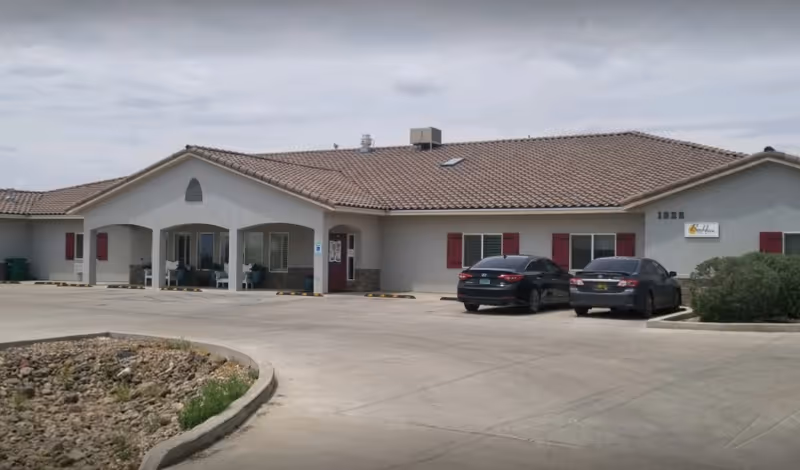 Exterior view of a single-story senior living facility building with a tiled roof, beige walls, and red window shutters. There is a covered entrance with seating and two parked cars in front. The building number 1928 is visible along with a sign that reads BeeHive Homes.