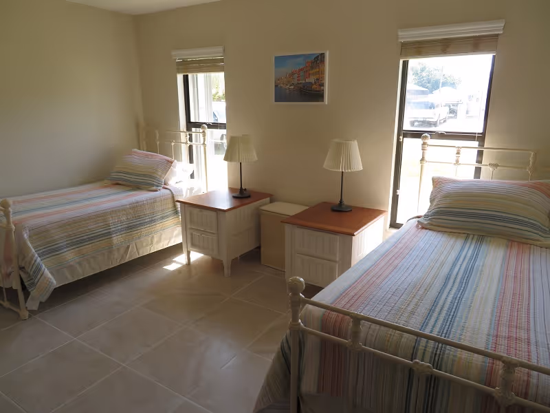 A bright bedroom with two single beds, each with a striped bedspread and pillow. Between the beds are two white nightstands with wooden tops, each holding a lamp with a pleated shade. Two windows with blinds allow natural light into the room. A framed picture hangs on the wall between the windows.