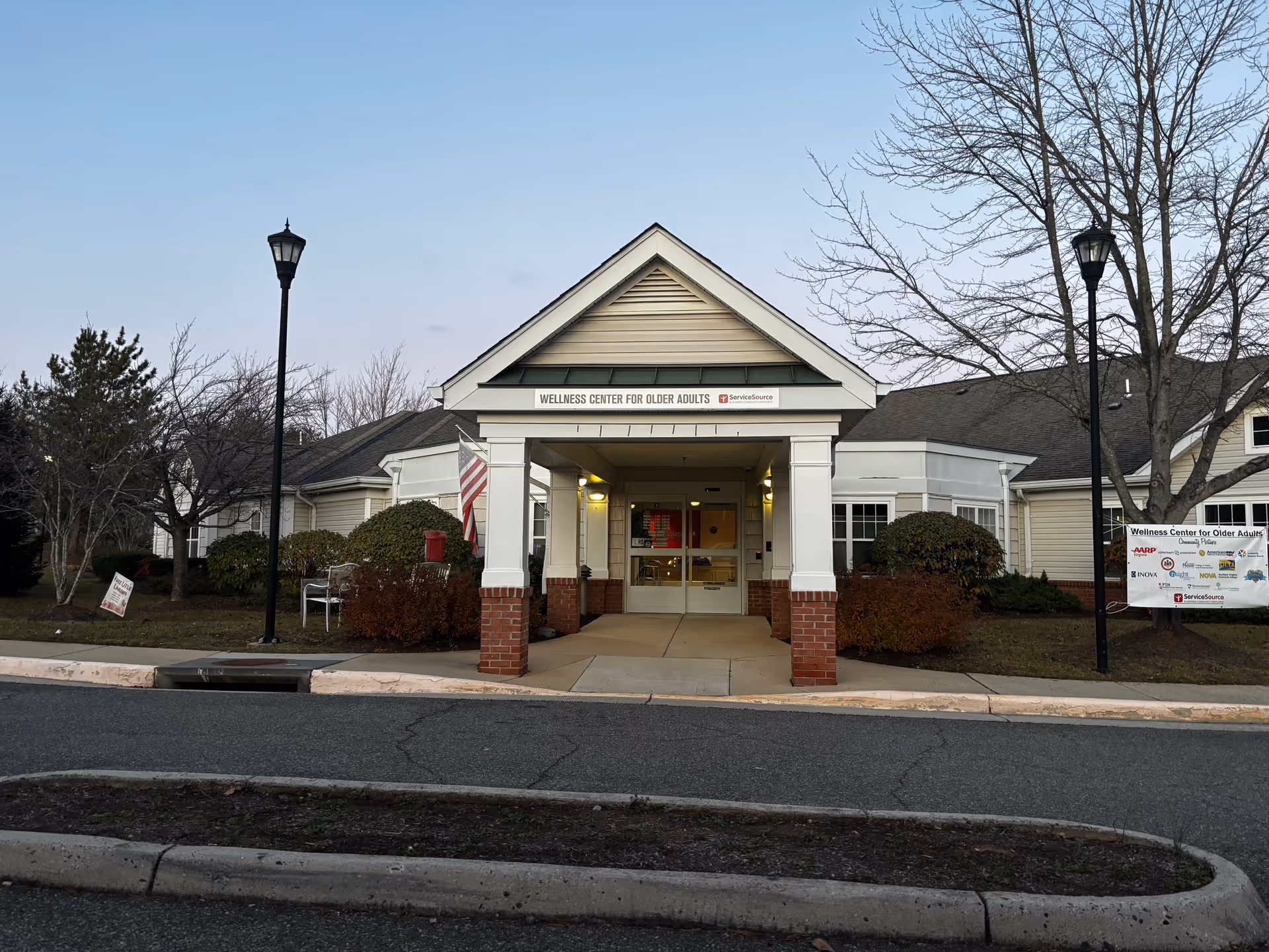 Exterior view of a single-story building with a covered entrance labeled 'Wellness Center for Older Adults'. The building has beige siding, white columns with brick bases, and an American flag near the entrance. There are leafless trees and bushes around the building, and two black street lamps on either side of the entrance.