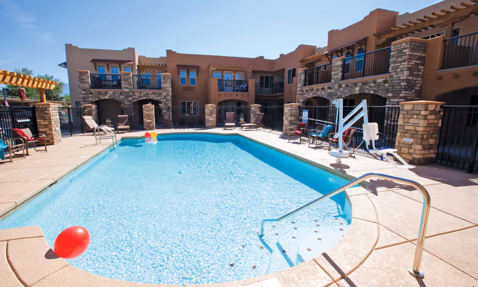 Outdoor swimming pool with clear blue water surrounded by a patio area with lounge chairs and a pool lift. The pool is located in the courtyard of a two-story building with balconies and stone accents under a clear blue sky.