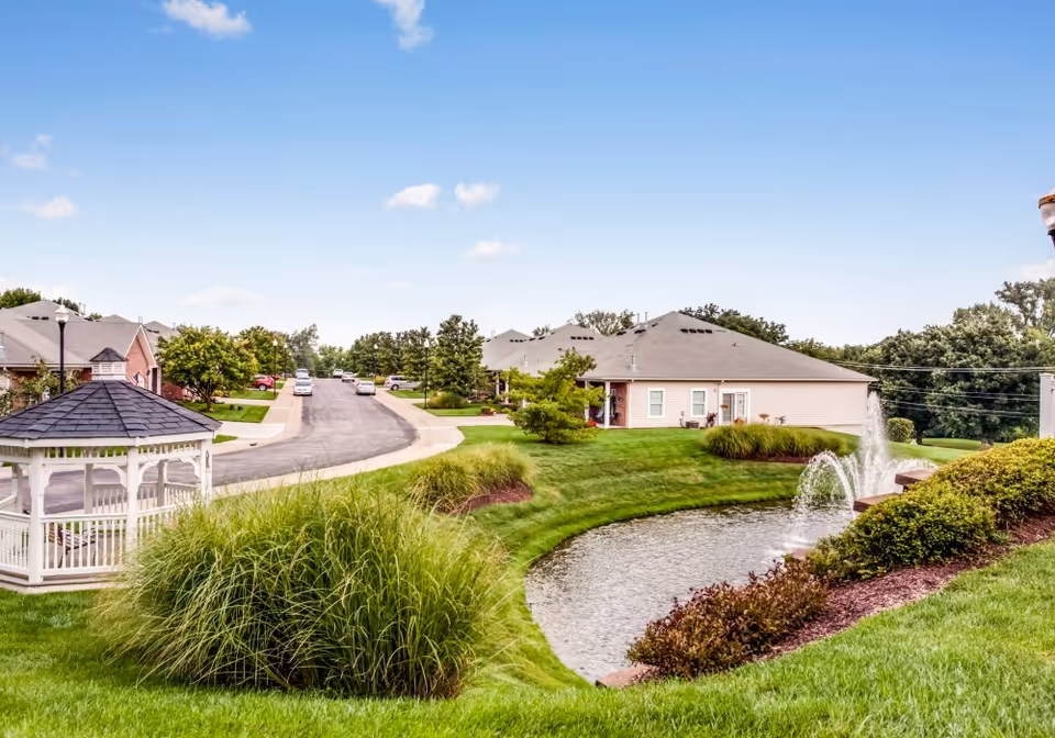 A peaceful outdoor scene at a senior living facility featuring a small pond with a fountain, well-maintained green lawns, bushes, and a white gazebo. Residential buildings and a street with parked cars are visible in the background under a clear blue sky.