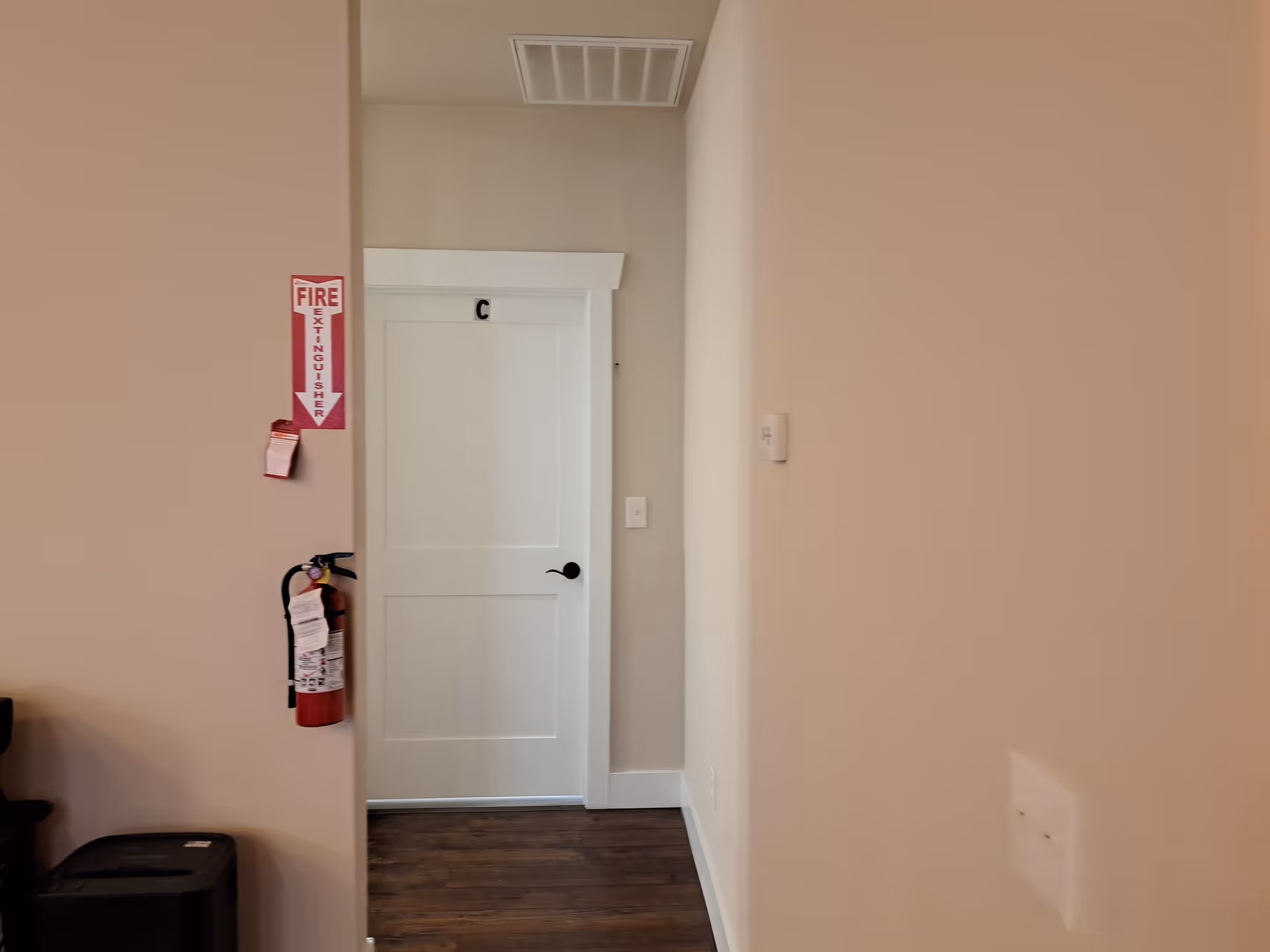Interior hallway view showing a white door labeled with the letter C, a fire extinguisher mounted on the wall with a red sign above it indicating its location, and wooden flooring.