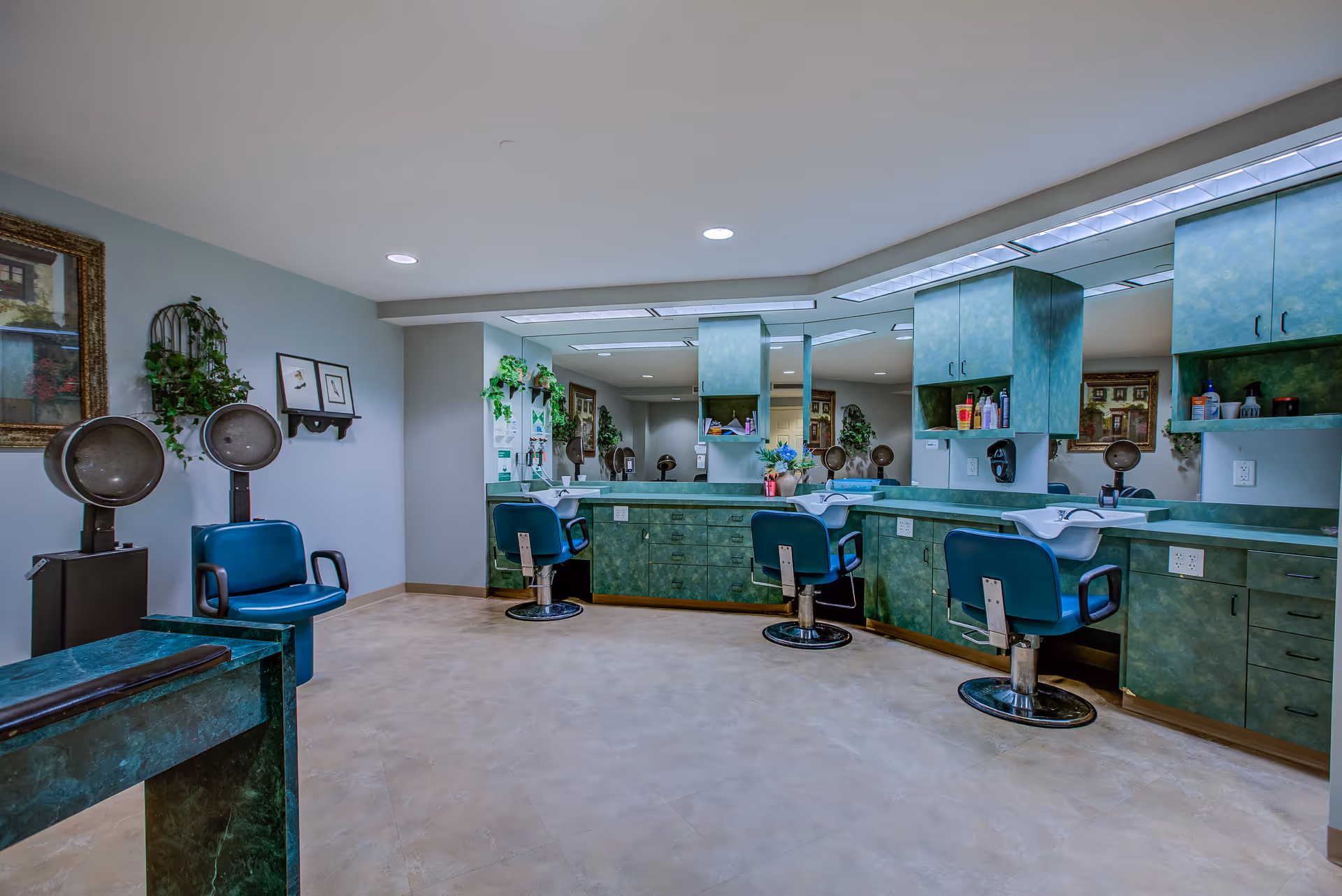 Interior view of a hair salon area in a senior living facility with green cabinetry, three blue salon chairs, hair washing sinks, hair dryers, and large mirrors on the walls. The room is well-lit with ceiling lights and decorated with framed pictures and plants.