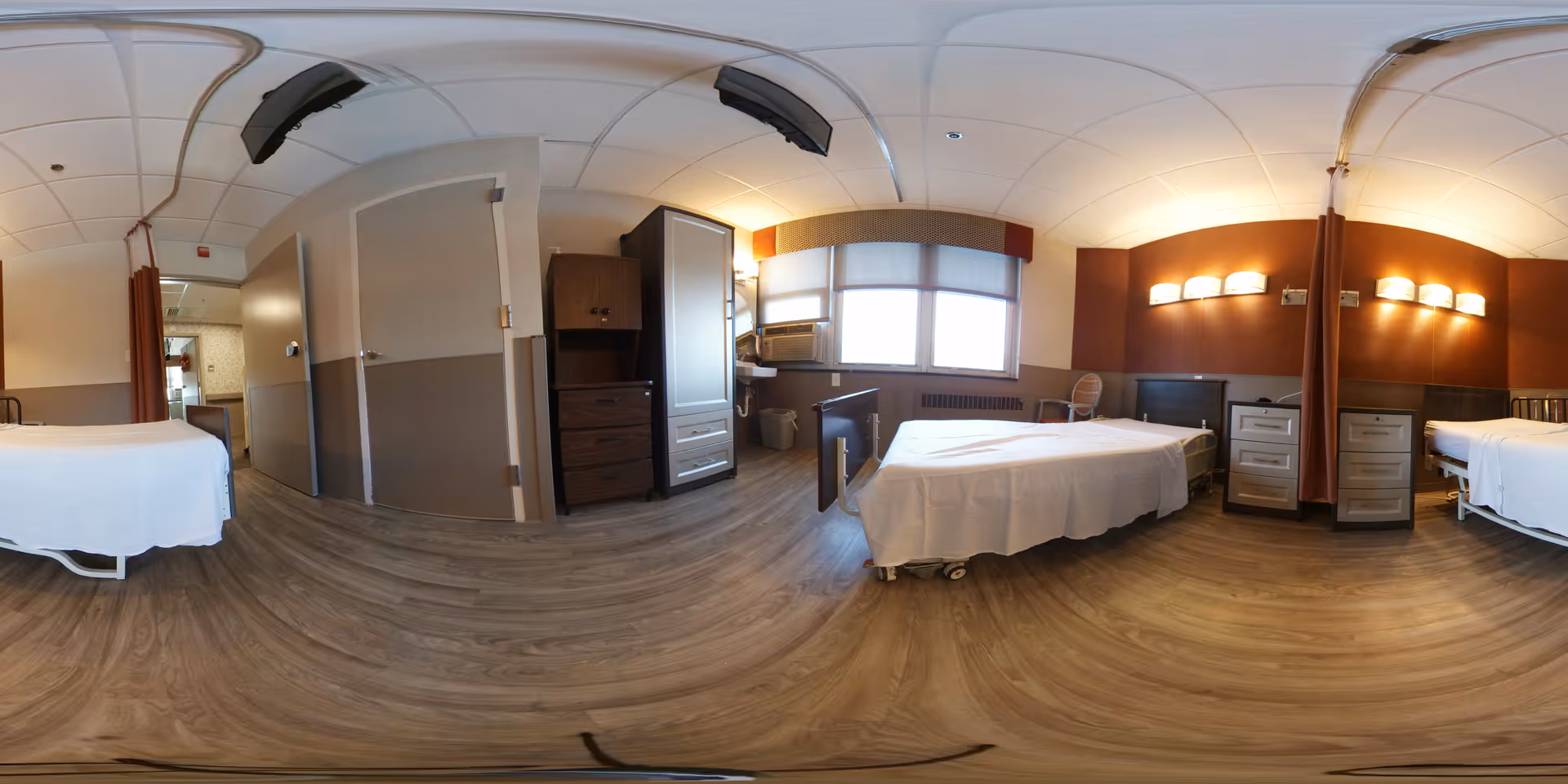 A wide panoramic view of a senior living facility bedroom with three hospital-style beds, each with white bedding. The room has wood flooring, beige and brown walls, and ceiling lights. There are bedside tables between the beds, a window with blinds, a wooden cabinet, and a chair near the window. Privacy curtains are partially visible between the beds.