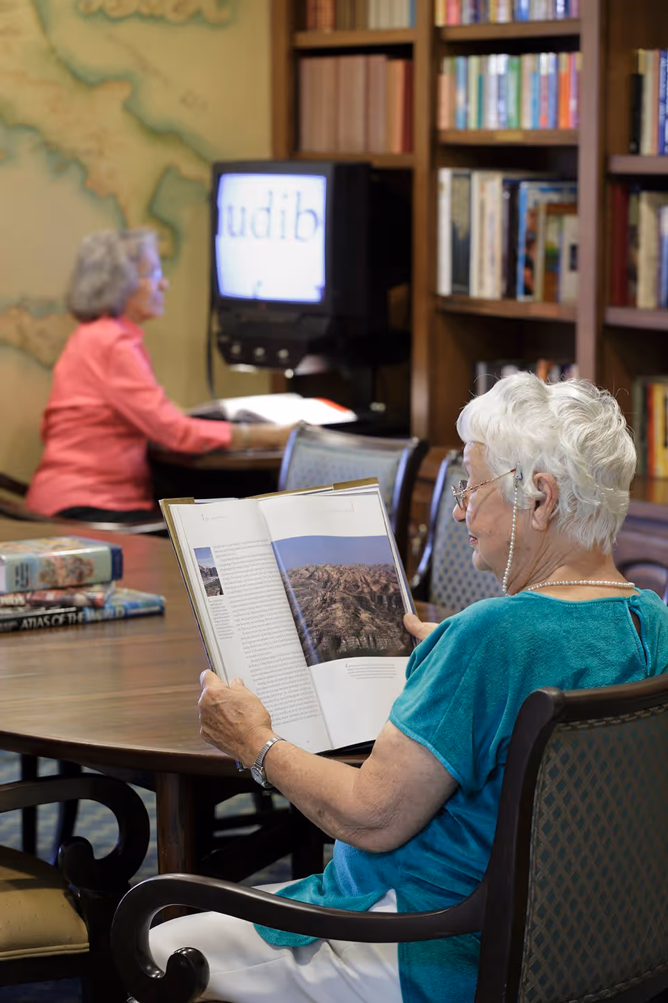 Two elderly women in a library or reading room. One woman with white hair and glasses is seated at a table reading a large book with a photo of a mountainous landscape. Another woman with gray hair and a pink top is seated at a desk in the background, looking at a screen. Bookshelves filled with books are visible behind them.