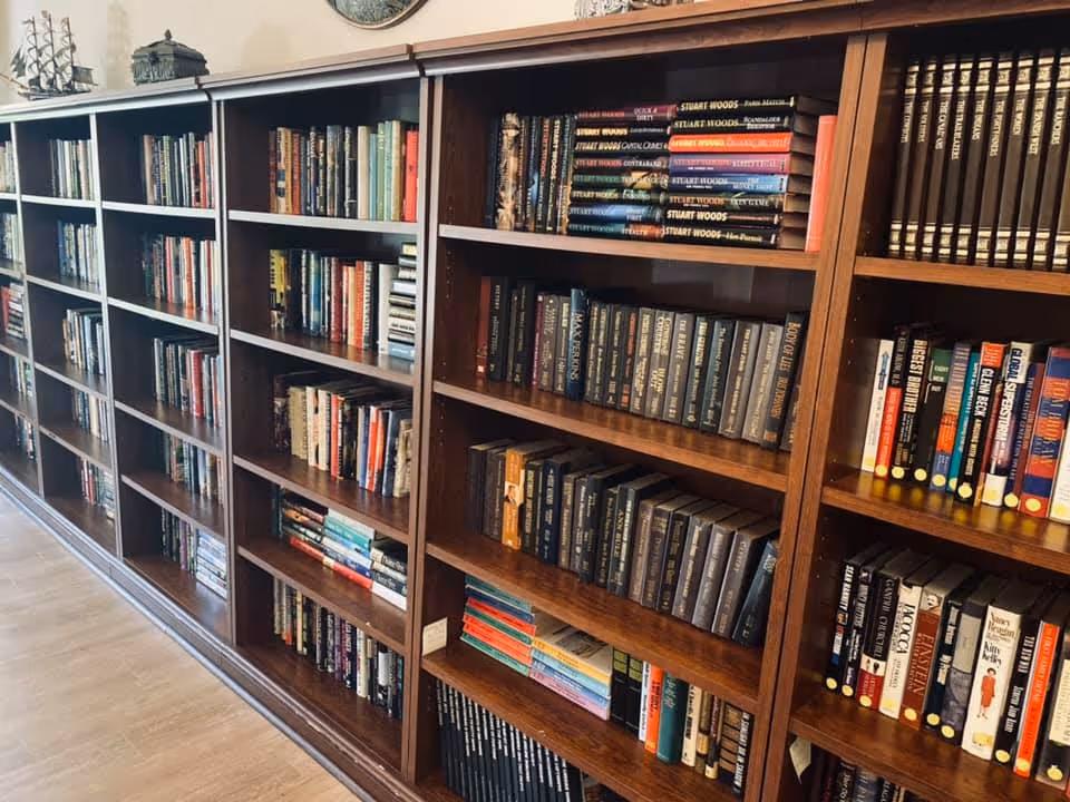 A long wooden bookshelf filled with a variety of books in a well-lit room with wooden flooring. Decorative items are placed on top of the bookshelf.