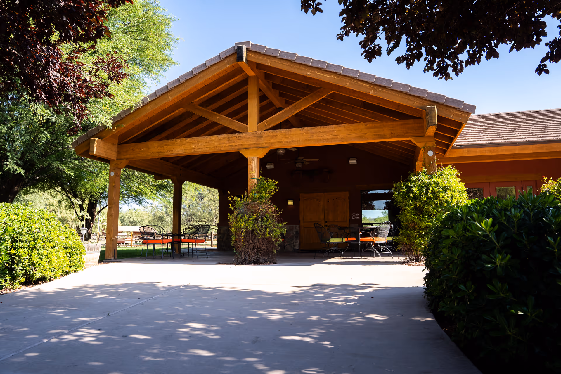 Outdoor covered patio area with wooden beams and roof, featuring several metal chairs with orange cushions and tables. The patio is surrounded by green bushes and trees under a clear blue sky.