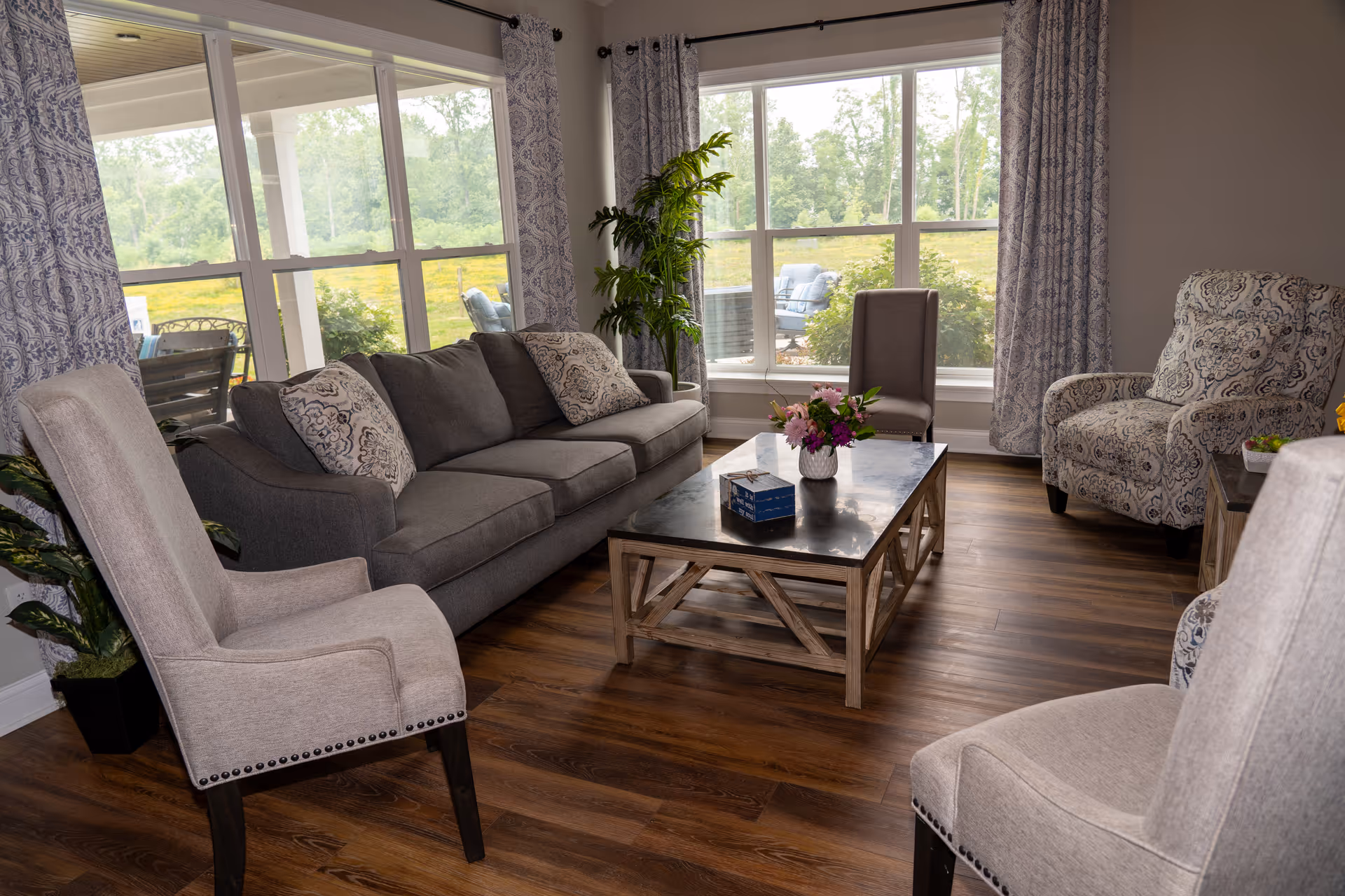 A cozy living room with a gray sofa adorned with patterned pillows, two beige upholstered chairs, and a patterned armchair. A wooden coffee table with a dark top holds a flower vase and a box of tissues. Large windows with patterned curtains let in natural light and offer a view of greenery outside. The room has wood flooring and some potted plants adding to the decor.