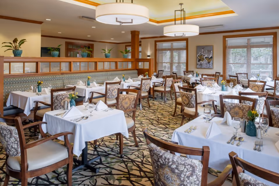 A well-lit dining room with multiple tables covered in white tablecloths, each set with folded napkins, glassware, and silverware. The chairs have patterned upholstery, and there are small flower arrangements in blue vases on each table. The room features large windows with blinds, decorative ceiling lights, and plants on a wooden partition.