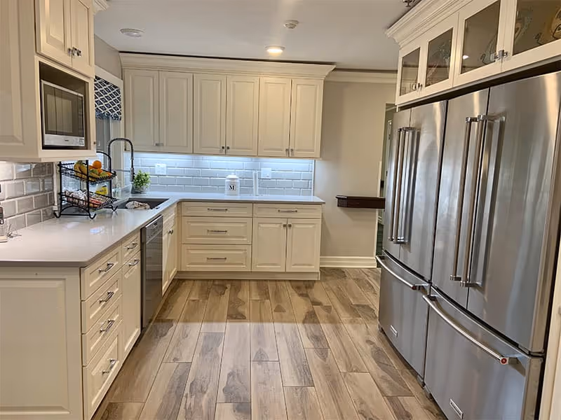 A modern kitchen with white cabinetry, stainless steel appliances including a double-door refrigerator and built-in microwave, a dishwasher, and a white subway tile backsplash. The floor is wood with a natural finish, and under-cabinet lighting illuminates the countertop.