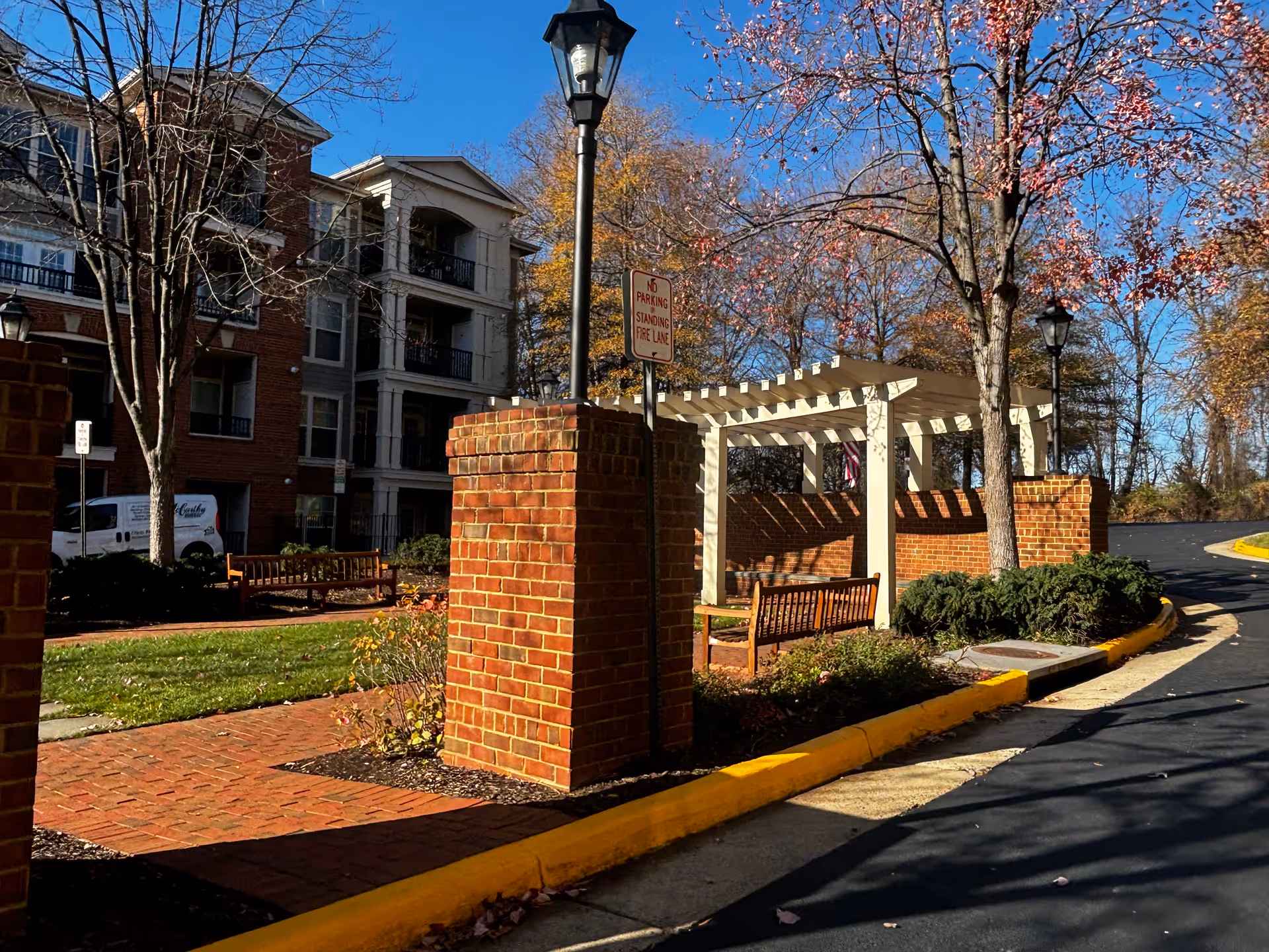 Exterior courtyard and entrance area of a senior living building with brick pillars, a white pergola, benches, trees, and a curved driveway.