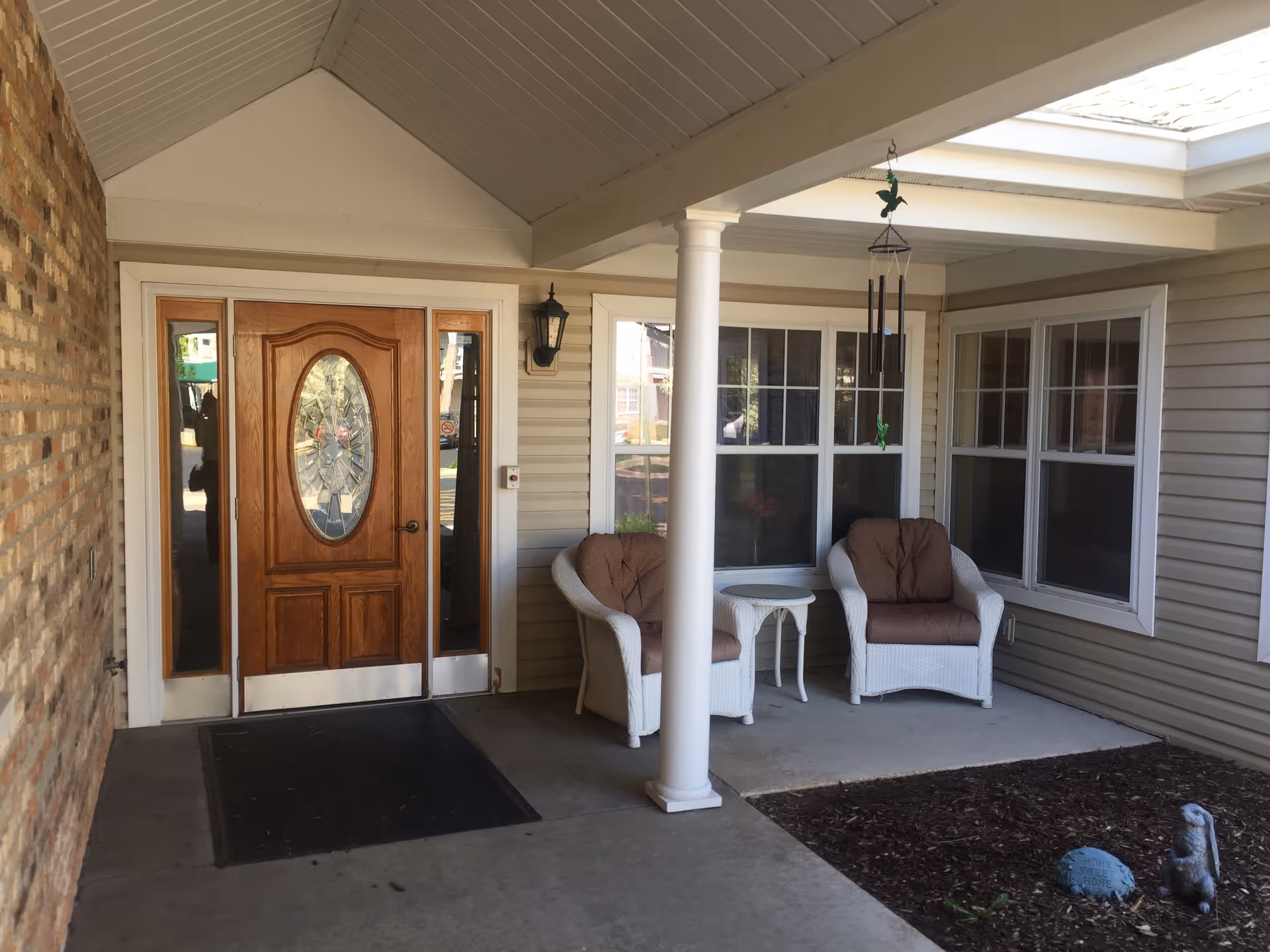 Covered outdoor entrance area with a wooden door featuring an oval glass panel, two white wicker chairs with brown cushions, a small white side table, a hanging wind chime, and a garden bed with a decorative stone and small statue.